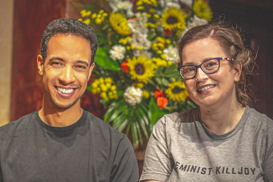 A black man wearing a dark top and a woman wearing glasses and a pale top, smiling at the camera with flowers in the background.
